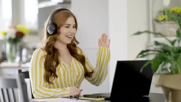Smiling woman with headphones waving goodbye at video call over laptop