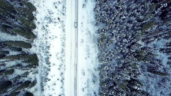 Aerial view looking down as a car drives along a winter road through pine forest alt