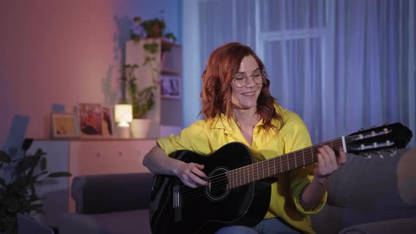 Woman with Glasses Learns Online Using Laptop to Play Guitar While Sitting on Couch at Home in alt