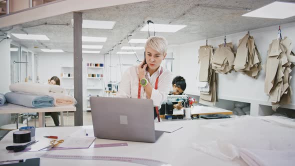 Female Fashion Designer Taking an Order By Cellphone Typing on Laptop Computer Standing at a Table alt