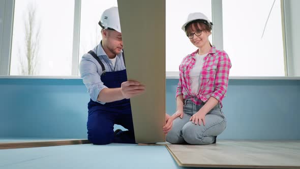 Man and Woman in Protective Helmets Check Locks of Laminate for Laying on Floor During Renovation alt