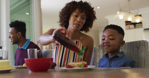 African american father smiling while pouring maple syrup on pancakes for her son at home alt