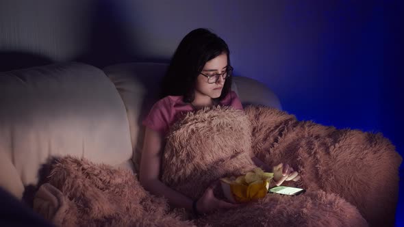 Girl Sitting on the Couch Watching Tv Uses Phone and Eating Chips in the Evening