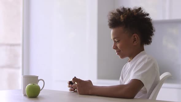 Cheerful African American Boy is Using Electronic Gadget at Table in Apartment Room Spbi alt