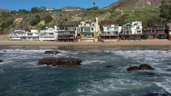 Aerial shot of ocean waves and cliff - waves washing up on a rock in sea and houses in the backgroun alt