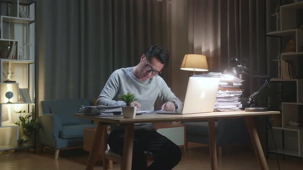 Asian Man Looking At Laptop And Taking Note While Working With Documents At Home alt