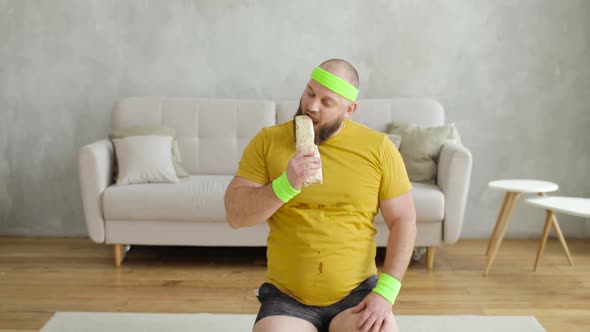 Fat Man Is Eating Shawarma Sitting on Fitness Mat at Home After Sport Exercises