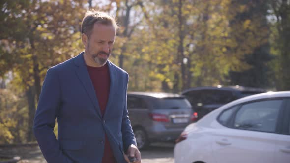 A Middleaged Caucasian Man Walks Toward His Car and Unlocks It with Remote Control in a Quaint Town