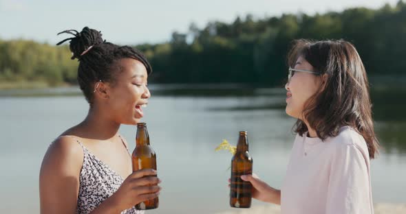 Two Beautiful Smiling Friends Spend Time Together on the Lakeshore Holding Glass Beer Bottles in alt