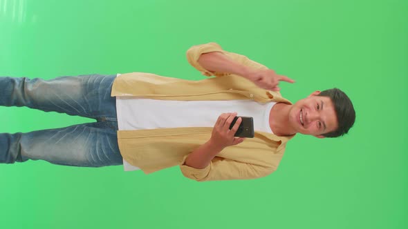 Asian Man Flipping Through The Mobile Phone With A Smile Pointing Up In Green Screen Studio alt