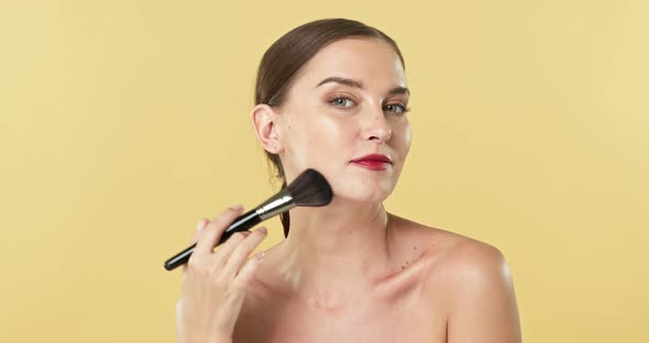 Young beautiful Caucasian woman using makeup brush on her face in isolated studio background