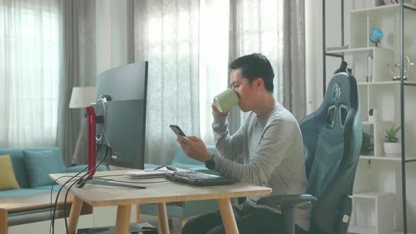 Asian Man Using Mobile Phone And Drinking Coffee While Using Desktop Computer For Working At Home alt