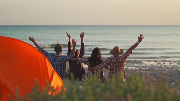 Rear View Four Best Friends Sitting at Sunset on Beach in Front of Sea and Raising Hands alt