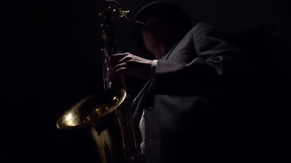 Male Saxophonist Dressed in a Classic Suit Blows a Saxophone in the Dark of Studio alt