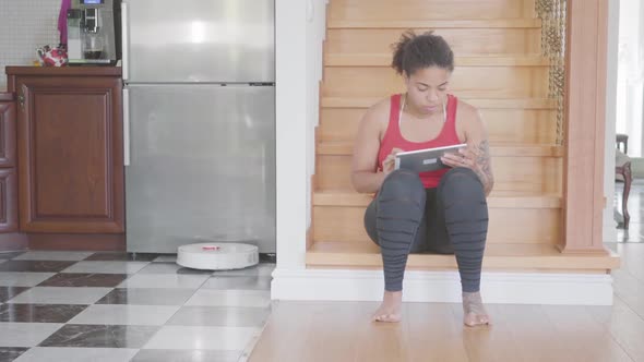 Cute African American Woman Sitting on the Stairs on the First Floor of Big House, Texting on Her alt