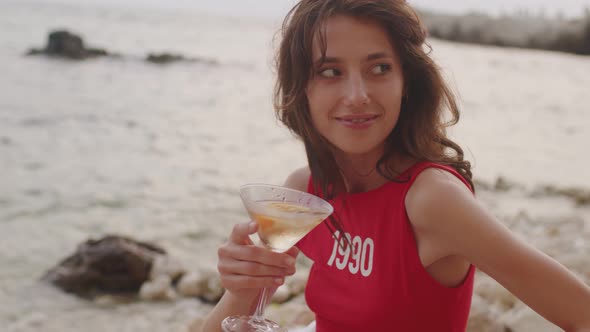 Young Brunette Woman in Red Shirt Sitting on Beach with Glass of Wine Enjoying Holidays