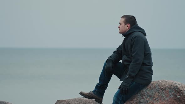 A young guy sits on a stone against the backdrop of the blue sea and thinks