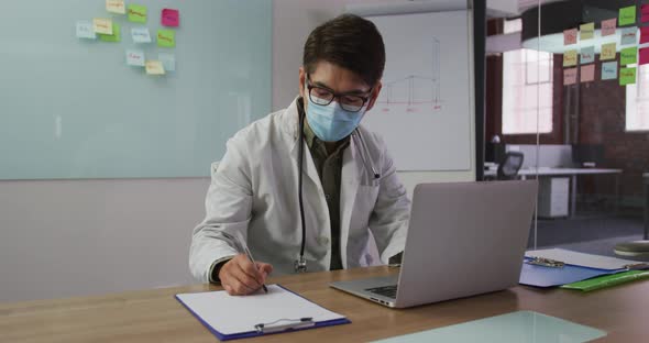 Asian male doctor wearing mask sitting at desk in meeting room making notes alt