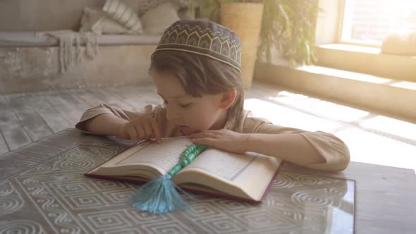 Adorable muslim child in prayer hat and arabic clothes with rosary beads reading holy Koran book 