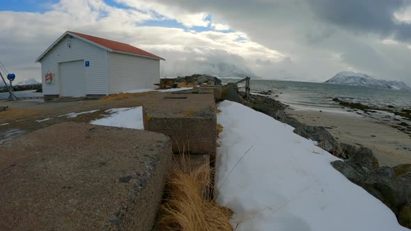 Low handheld dolly shot along breakwater wall with thick snow, dark forboding clouds and mountains i alt