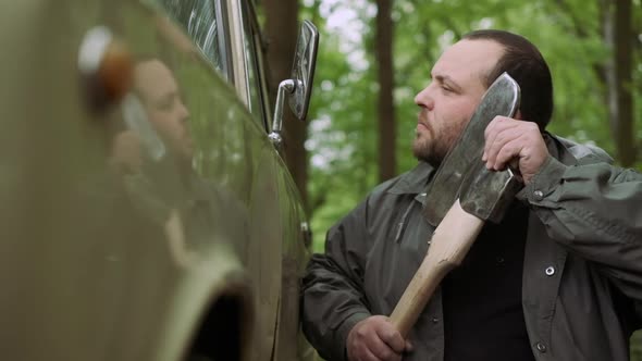 Slow Motion Of Casual Strong Man Has Shave with Ax Near Classic Car in Wild Deciduous Forest. alt