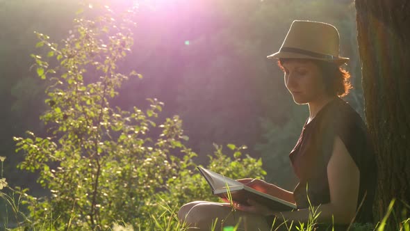 A Young Girl in a Straw Hat and Dress Sits Under a Tree, Looks Around, Then Reads a Book alt