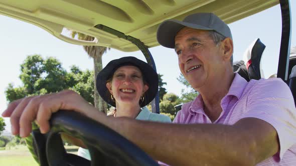Caucasian senior couple driving a golf cart with clubs on the back at golf course alt