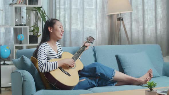 Full View Of Asian Woman Relaxing Leaning On Sofa To Sing And Play A Guitar At Home alt