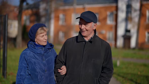 an Elderly Couple Walks Arm in Arm Against the Background of Blurred Lowrise Houses alt