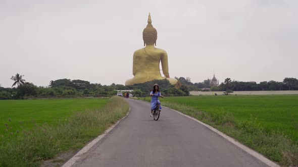 Asian woman cycling a bicycle near the Giant Golden Buddha in Wat Muang in Ang Thong alt