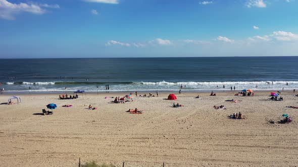 A drone view of people relaxing on the beach on a sunny day. The drone camera slowly booms up & tilt alt