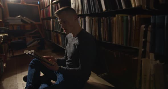 A Young Guy Is Sitting on the Floor Between the Bookshelves and Leafing Through a Book.  alt