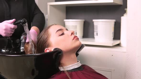 A Gloved Hairdresser Washes the Hair of a Blonde Client in a Black Washbasin