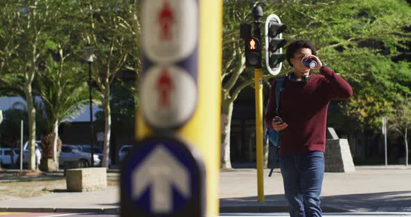 Happy african american man using smartphone, drinking coffee, crossing street wearing backpack alt