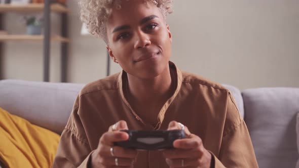 African Woman Player Sit on Couch Cross Legged at Home Holding Console Game Pad alt