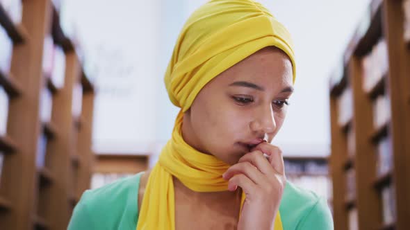 Asian female student wearing a yellow hijab sitting with an open book and using laptop alt