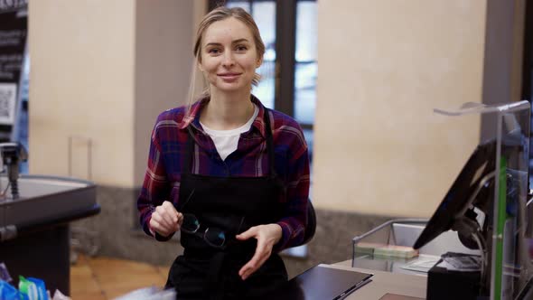 Woman Putting on Eyeglasses Working at the Checkout in a Supermarket alt