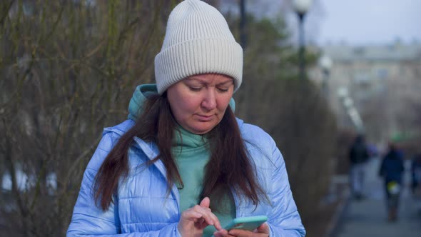 Woman Texting Message In Smartphone Standing On Street In City Park Near Trees
