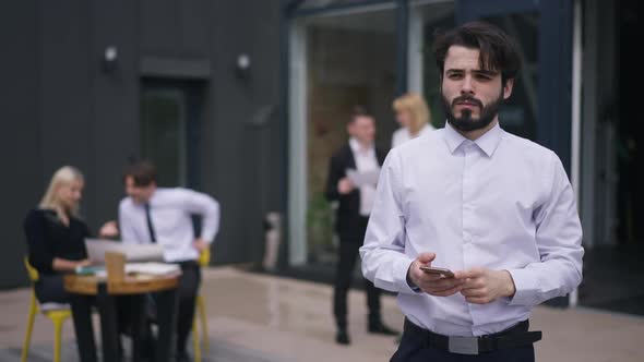 Portrait of Handsome Caucasian Bearded Young Man Standing on Office Terrace with Smartphone Thinking alt