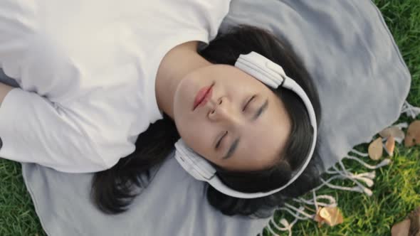 Asian female wearing headphones listening to music while lying on the floor grass outdoors.