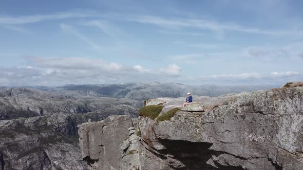 Aerial Shot of a Female Hiker Wearing Blue Jacket Sitting at the Edge of a Cliff alt