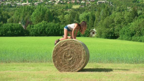 A beautiful brunette woman playing trying to roll a large bale of hay on a farm in France. alt