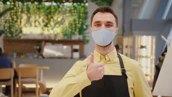 Male Waiter Cafe Owner Wearing Mask and Gloves Stands and Shows Thumb alt