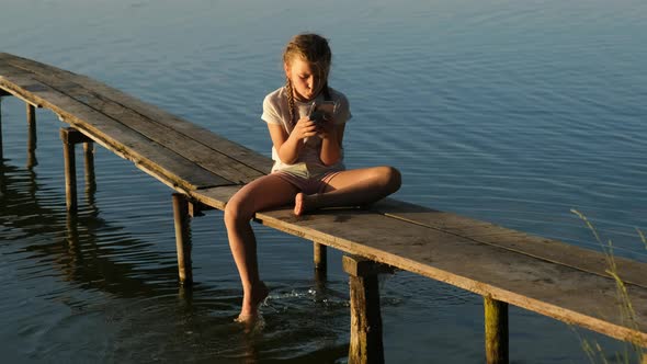 A little girl with a smartphone sits on a wooden pier on the lake during sunset.