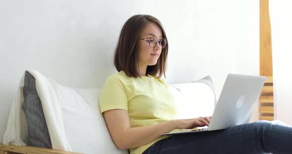 Young Beautiful Woman is Sitting in a Large White Armchair and Chatting Using a Laptop