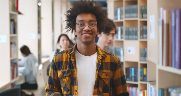 Confident Handsome African Student Smiling at Camera with Library Bookshelves on Background alt