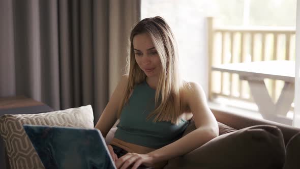Blonde Woman Sitting on Sofa in Country House Browsing Social Media on Laptop alt