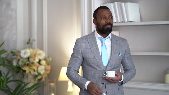 a Bearded AfricanAmerican Man in a Plaid Suit with a Blue Tie Stands Thoughtfully with a Coffee Cup alt