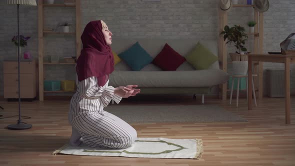 Muslim Woman in a Traditional Scarf Praying on Rug in Her Living Room alt