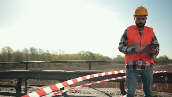 Young Bearded Man Inspector in an Orange Vest and Protective Helmet with Tablet alt